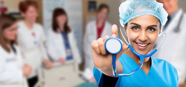 Smiling nurse in blue scrubs holding a stethoscope with medical staff blurred in the background